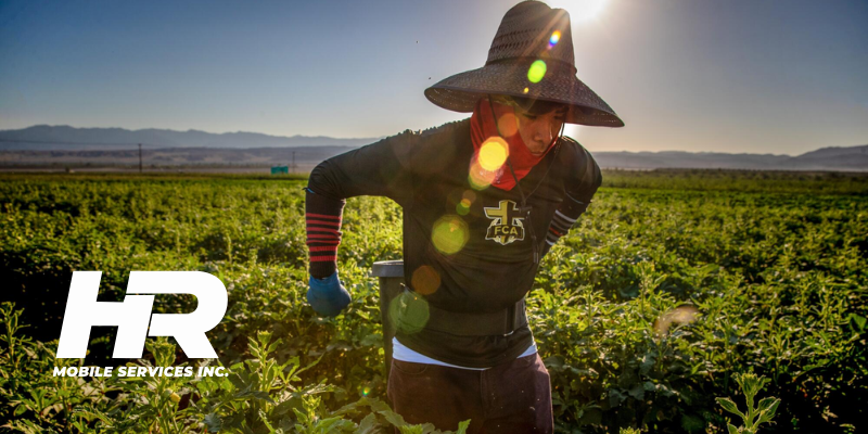 Web Banners - News Blogs 4 - The AG Center Farmer wearing a wide-brim hat and gloves harvesting crops in a sunlit field, with the HR Mobile Services Inc. logo in the bottom-left.